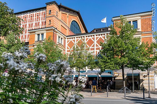 Gebäude mit Fachwerkfassade, Bäumen und Menschen vor dem Eingang bei klarem Himmel.