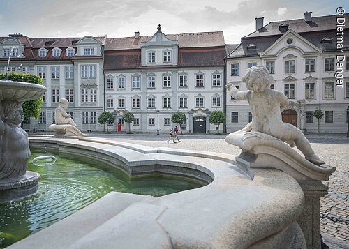 Brunnen mit Puttenfiguren im Vordergrund und historischen Gebäuden im Hintergrund auf einem gepflasterten Platz.