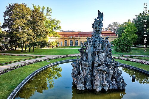 Brunnen mit Figuren in einem runden Wasserbecken vor einem gelben Gebäude und Park mit Bäumen und Blumenbeeten.