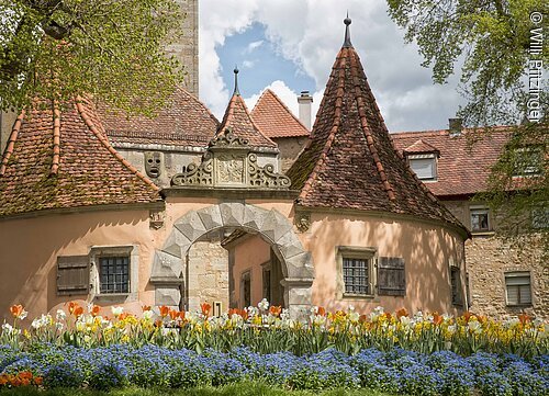 Rundturm mit Spitzdach und Blumenbeet aus Tulpen und Vergissmeinnicht im Vordergrund bei bewölktem Himmel.