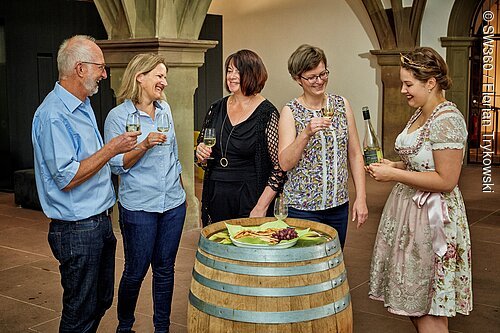 Fünf Personen stehen um ein Fass mit Snacks, trinken Wein und unterhalten sich in einem Innenraum.