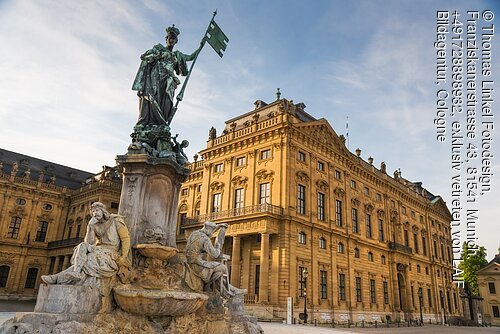Brunnen mit mehreren Figuren vor einem großen barocken Gebäude unter blauem Himmel.