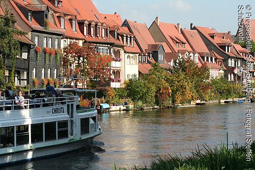 Fluss mit Ausflugsboot und Fachwerkhäusern mit roten Dächern und Bäumen am Ufer im Sonnenschein