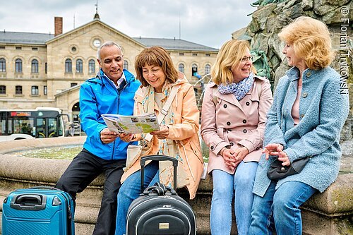 Vier Menschen mit Koffern sitzen an einem Brunnen und schauen auf eine Karte vor einem historischen Gebäude.