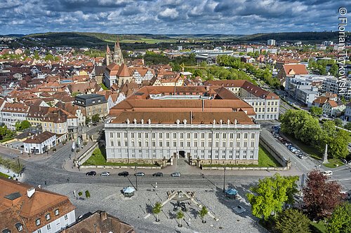 Luftaufnahme eines historischen Gebäudes mit rotem Dach in einer Stadt mit vielen Häusern und bewölktem Himmel.