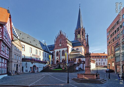 Stiftsplatz (Aschaffenburg, Spessart-Mainland) Marktplatz mit Kopfsteinpflaster, historischem Fachwerkhaus, Kirche mit Turm und modernem Gebäude bei klarem Himmel