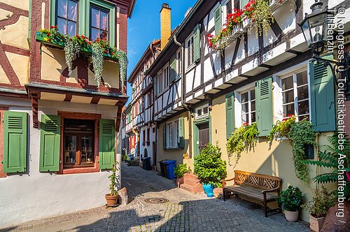 In der Altstadt (Aschaffenburg, Spessart-Mainland) Enge gepflasterte Gasse mit Fachwerkhäusern, grünen Fensterläden, Blumen und einer Holzbank bei Sonnenschein.