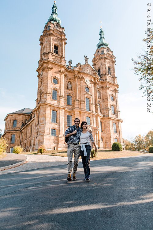 Basilika Vierzehnheiligen (Bad Staffelstein, Obermain.Jura) Paar mit Rucksack geht vor einer großen historischen Kirche mit zwei Türmen bei Tageslicht spazieren.