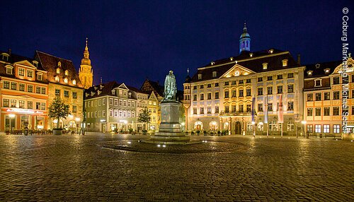 Beleuchteter Marktplatz mit Statue und historischen Gebäuden bei Nacht, Kopfsteinpflaster sichtbar.