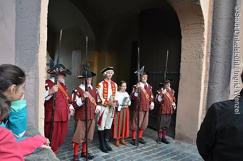 Sechs Personen in historischen Kostümen mit Hellebarden und Musikinstrumenten vor einem Tor, zwei Zuschauer im Vordergrund.