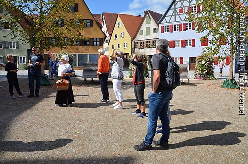 Gruppe von Menschen steht auf gepflastertem Platz vor bunten Fachwerkhäusern, eine Frau trägt historische Kleidung.
