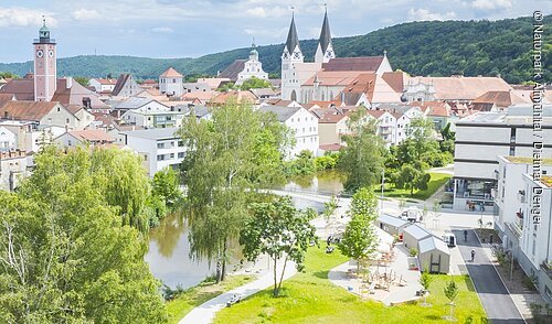 Wanderer an der Altmühl (Eichstätt, Naturpark Altmühltal) Stadtansicht mit Fluss, Bäumen, Kirche mit zwei Türmen und Wohngebäuden bei bewölktem Himmel.