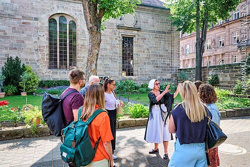 Kostümführung (Erlangen, Städteregion Nürnberg) Frau in historischer Kleidung erklärt sieben Personen im Innenhof vor einer Kirche etwas.