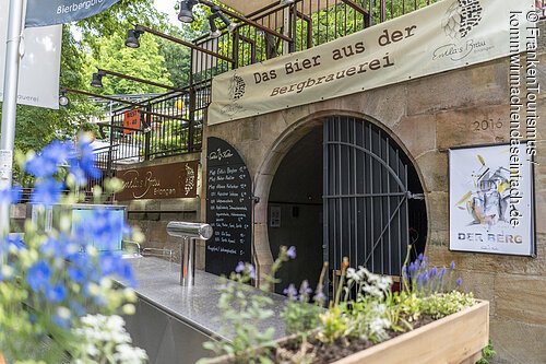 Entla’s Keller am Burgberg (Erlangen, Städteregion Nürnberg) Außenansicht eines Biergartens mit Zapfanlage, Blumen im Vordergrund und Eingang zu einem Keller mit Banner „Das Bier aus der Bergbrauerei“.