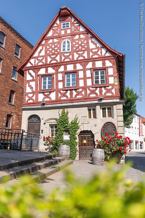 Fachwerkhaus mit roten Balken, weißen Wänden, zwei Holztüren, zwei Weinfässern und roten Blumen am Eingang.