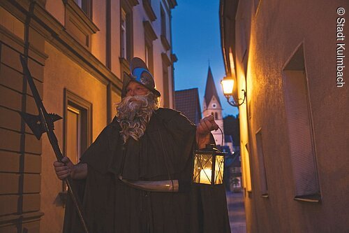 Person mit Bart und Helm hält Laterne und Hellebarde in einer engen Gasse bei Abenddämmerung