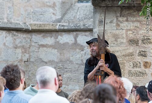 Mann in historischem Kostüm mit Hellebarde und Laterne spricht vor einer Menschenmenge vor einer Steinmauer.