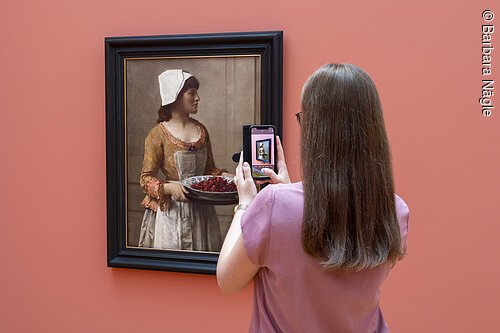 Frau fotografiert Gemälde einer Frau mit Haube und Schale voller Kirschen in Museum vor roter Wand.