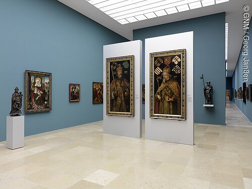 Museumsgalerie mit blauen Wänden, zwei großen mittelalterlichen Gemälden auf weißen Stellwänden und Skulpturen auf Podesten.