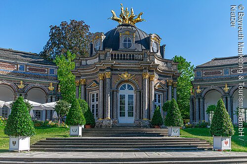 Barocker Pavillon mit goldener Skulptur auf der Kuppel, umgeben von grünen Büschen und blauem Himmel.