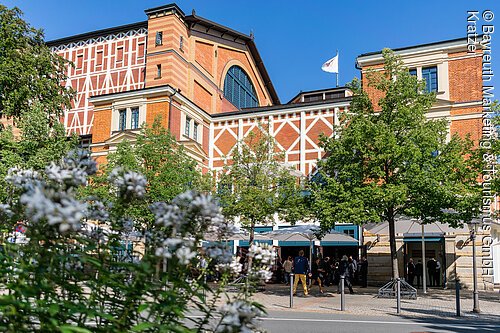 Gebäude mit Fachwerkfassade, Bäumen und Menschen vor dem Eingang bei klarem Himmel.