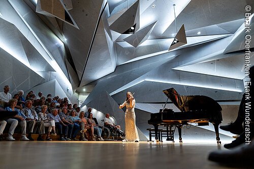 Eine junge Frau im Abendkleid spielt Violine vor einem geometrisch anmutenden Hintergrund. Neben ihr ein Flügel sowie Publikum.