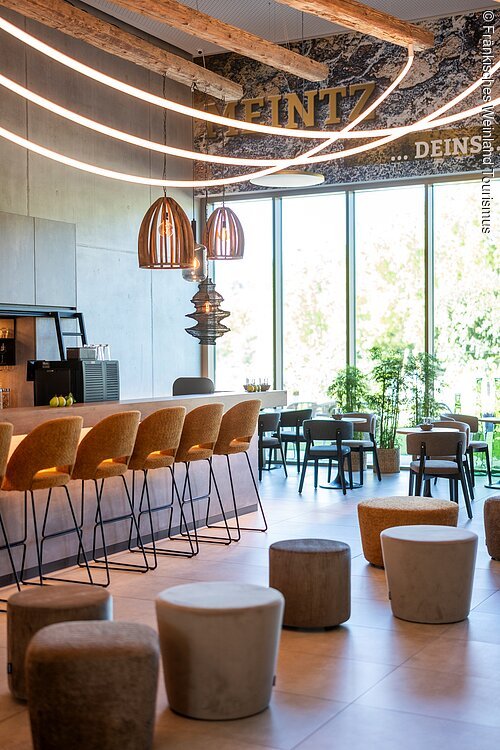 Moderner Gastraum mit Barhockern, Sitzhockern, großen Fenstern und hängenden Lampen unter Holzdeckenbalken.