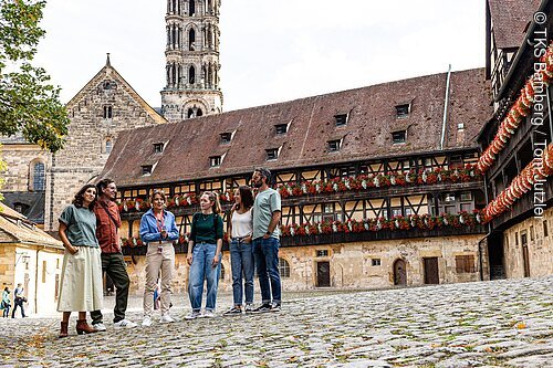 Sechs Personen stehen auf einem gepflasterten Platz vor einem Fachwerkhaus mit Blumenschmuck und einem Kirchturm im Hintergrund.