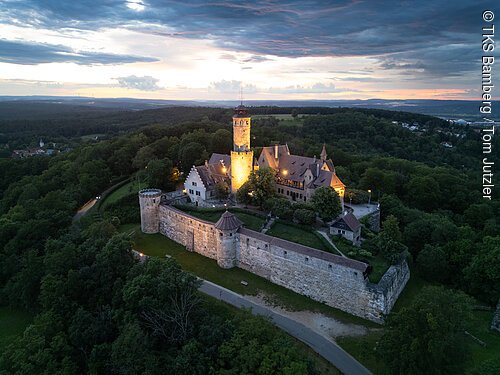 Luftaufnahme einer beleuchteten Burg mit Wehrmauer umgeben von Wald bei Sonnenuntergang.