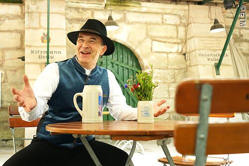 Mann mit Hut und Weste sitzt an Holztisch mit Bierkrug und Vase mit Blumen in Biergarten.