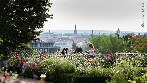 Drei Personen liegen auf einer Mauer in einem Park mit Blumen und Blick auf eine Stadt mit Kirchtürmen im Hintergrund.