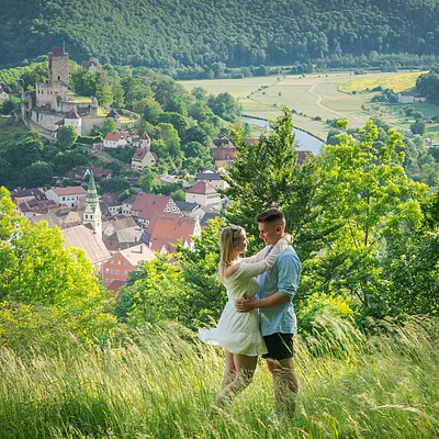 Paar umarmt sich auf einer Wiese mit Blick auf eine Stadt und eine Burg im Hintergrund.