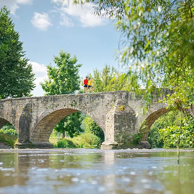 Steinbrücke mit drei Bögen über Fluss, umgeben von Bäumen, zwei Personen stehen oben auf der Brücke.