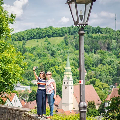 Zwei Frauen machen Selfie vor grünem Hügel und Kirche mit spitzem Turm, Laterne im Vordergrund.