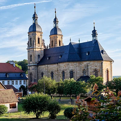 Kirche mit zwei Türmen und umliegenden Häusern bei klarem Himmel und Bäumen im Vordergrund.