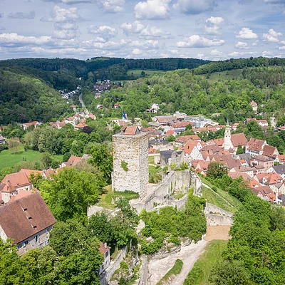 Luftaufnahme einer Burg mit Turm und umliegendem Dorf in einer grünen Hügellandschaft unter bewölktem Himmel.