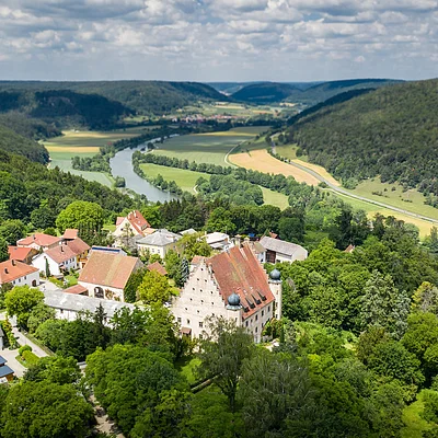 Luftaufnahme eines Dorfes mit Fluss, umgeben von Wäldern und Hügeln unter bewölktem Himmel.