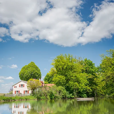 Haus mit roten Fensterläden am Ufer eines Sees, umgeben von grünen Bäumen und blauem Himmel mit Wolken