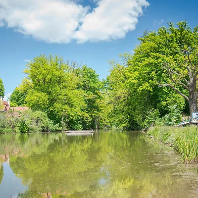 Fluss mit grünen Bäumen, zwei Häusern links und rechts und Personen am Ufer unter blauem Himmel