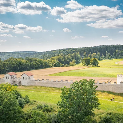 Landschaft mit Wiesen, Feldern, Bäumen und einer historischen Steinmauer mit Türmen unter bewölktem Himmel.