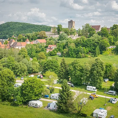 Blick auf einen Campingplatz mit Wohnmobilen vor bewaldeten Hügeln und einer Burg auf einem Hügel im Hintergrund.