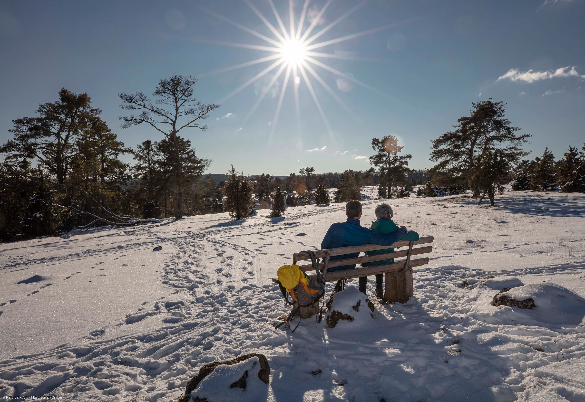 Zwei Personen sitzen auf einer Bank in verschneiter Landschaft unter strahlender Sonne am blauen Himmel.