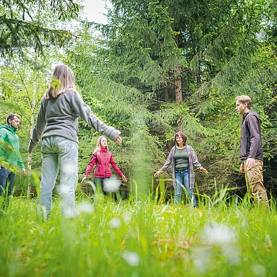 Fünf Personen stehen mit Abstand im Kreis auf einer Wiese vor Nadelbäumen im Wald.