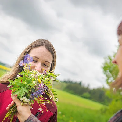 Frau riecht an einem Strauß mit Kräutern, der ihr von einer anderen Person im Freien überreicht wird.