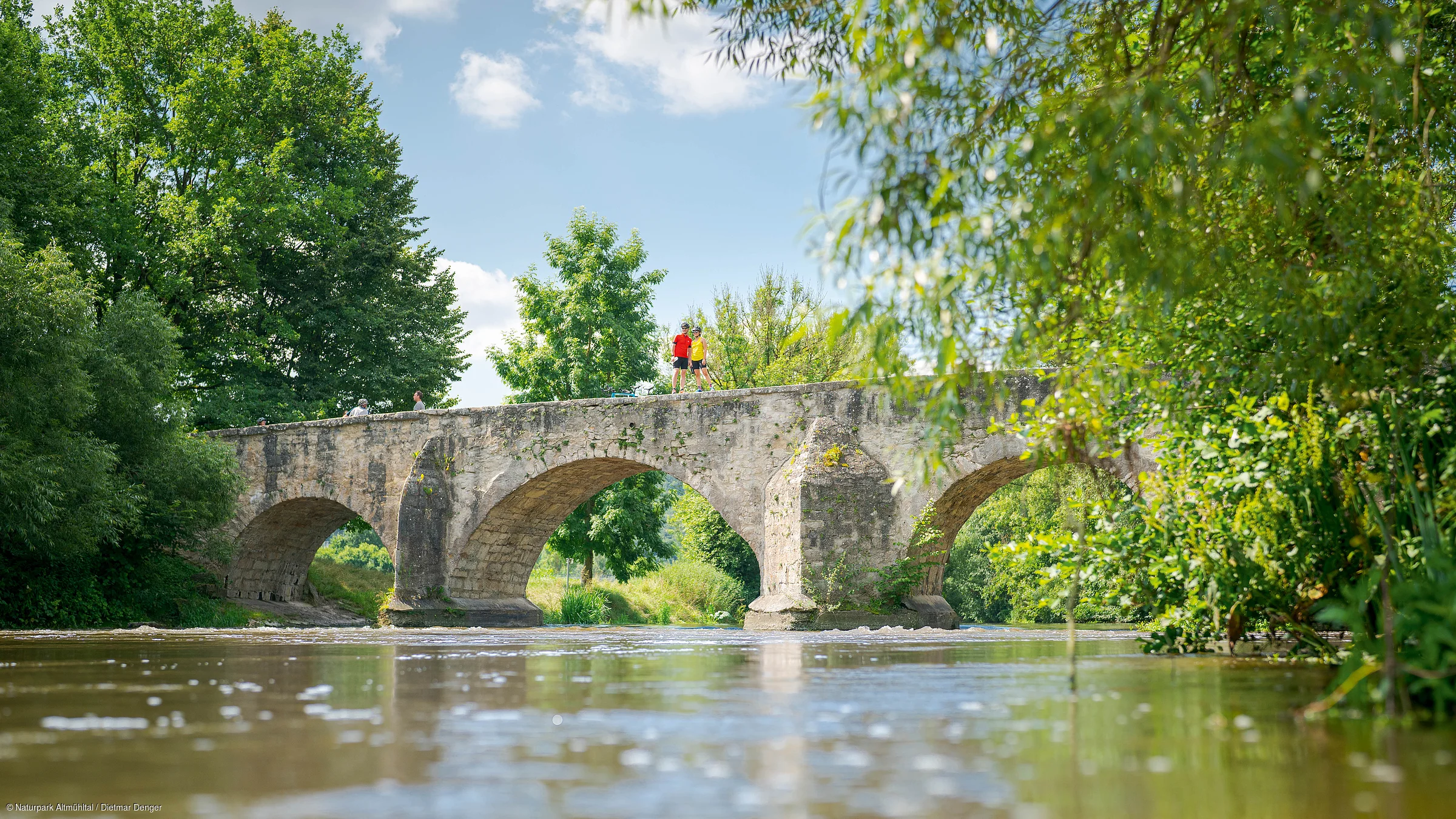 Steinbrücke mit drei Bögen über Fluss, umgeben von Bäumen, zwei Personen stehen oben auf der Brücke.