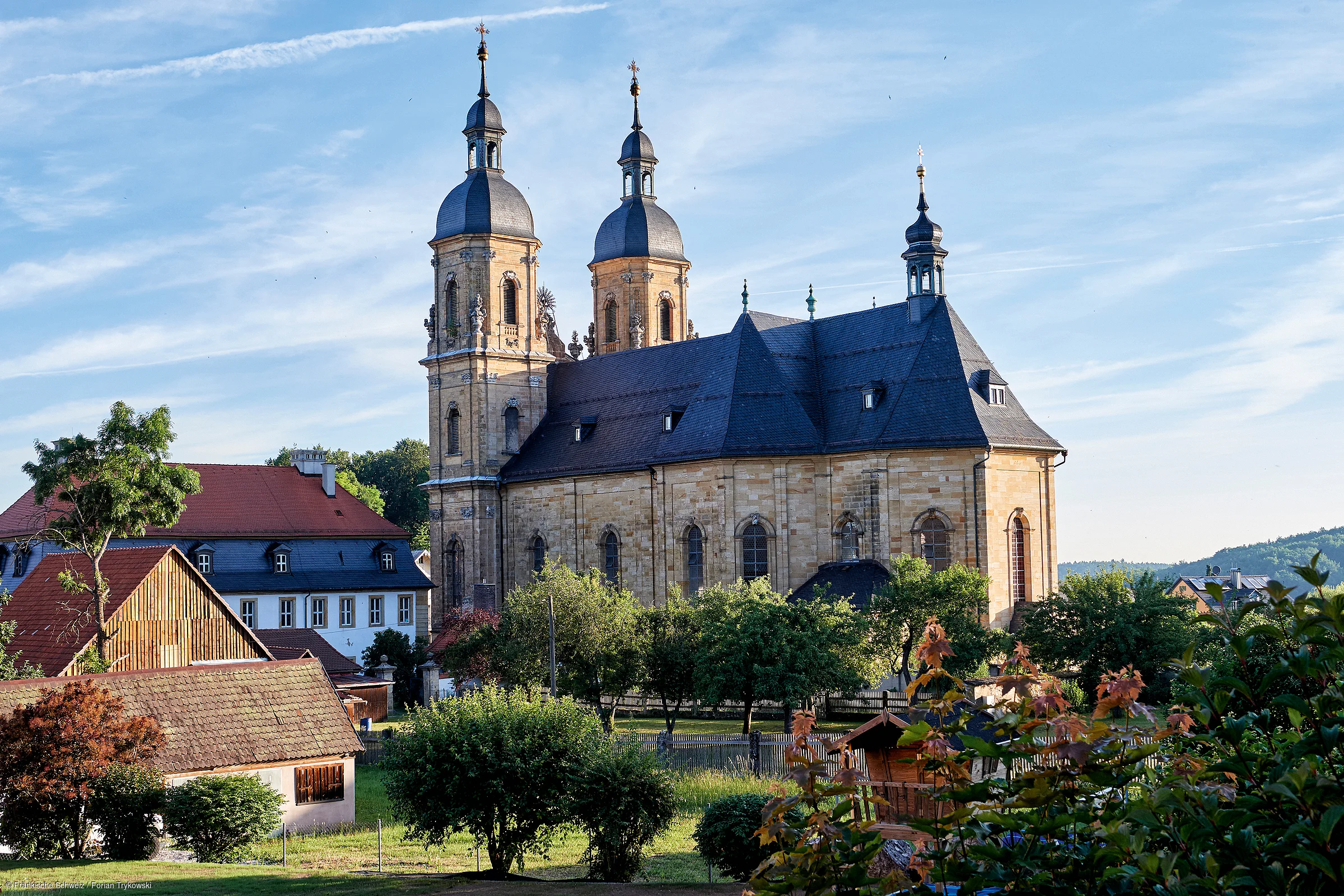 Kirche mit zwei Türmen und umliegenden Häusern bei klarem Himmel und Bäumen im Vordergrund.
