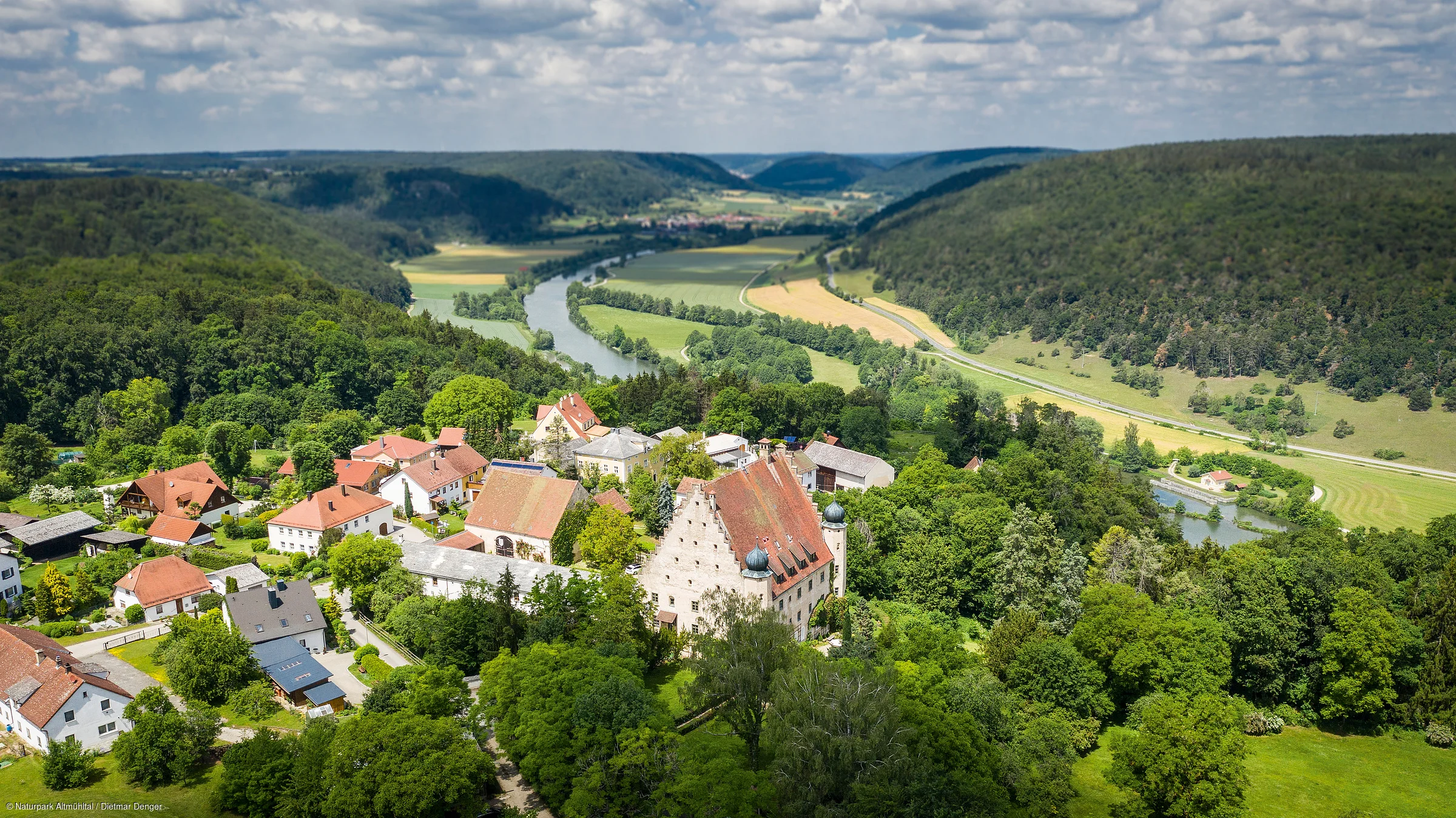 Luftaufnahme eines Dorfes mit Fluss, umgeben von Wäldern und Hügeln unter bewölktem Himmel.