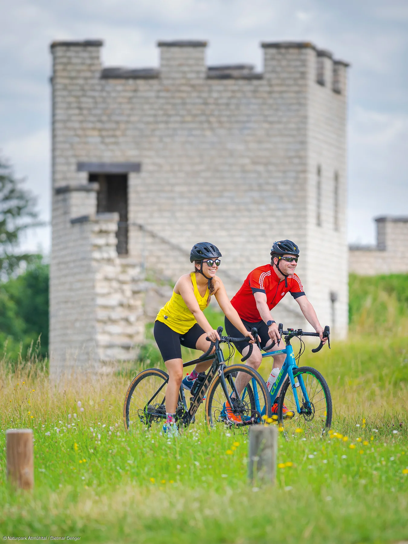Zwei Radfahrer mit Helm und Sportkleidung fahren auf einem Weg vor einem rekonstruierten römischen Kastell Grünen.