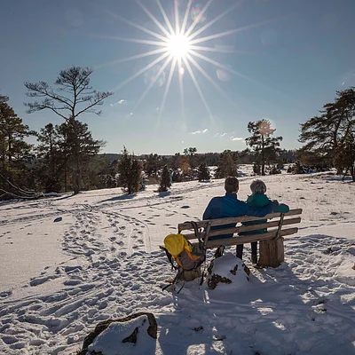 Zwei Personen sitzen auf einer Bank in verschneiter Landschaft unter strahlender Sonne am blauen Himmel.