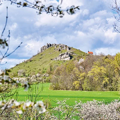 Grüner Berg mit Felsen und Kapelle, umgeben von blühenden Bäumen und bewölktem Himmel.